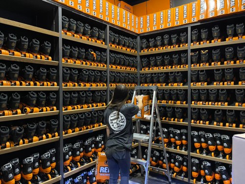 A person on a ladder organizes orange and black boots on shelves in a retail store.