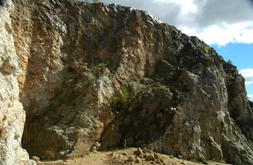 A rugged rock face with a steep cliff, surrounded by sparse vegetation and a cloudy sky above.