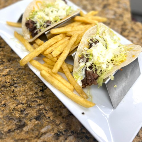 Two tacos with beef and lettuce served in metal holders, accompanied by a side of golden French fries on a white plate.