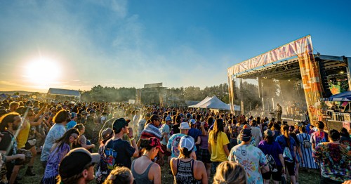 A large crowd enjoys a music festival at sunset, with a stage and colorful decorations in the background.