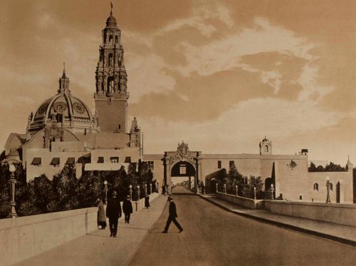 Historic sepia-toned image of a grand building with a tower, featuring a bridge and pedestrians walking.