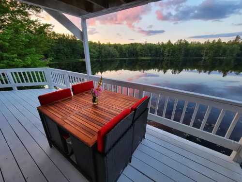 A wooden table with red cushions on a deck overlooking a calm lake at sunset, surrounded by trees.