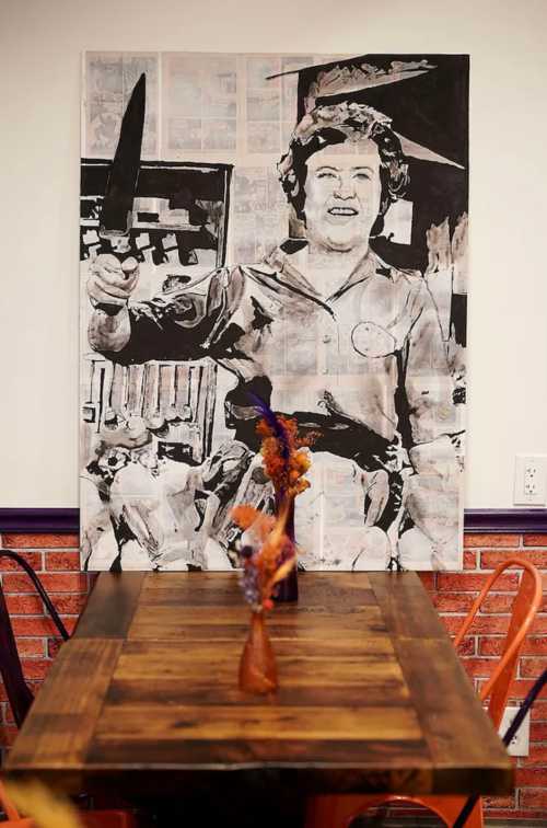 A black and white mural of a smiling woman holding a knife, displayed above a wooden table with flowers.