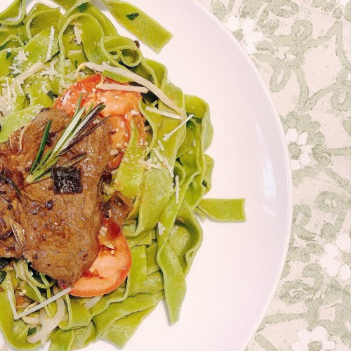 A plate of green fettuccine topped with sliced tomatoes, herbs, and a piece of meat, garnished with cheese.