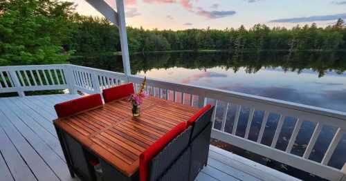 A wooden table with red chairs on a deck overlooking a calm lake surrounded by trees at sunset.