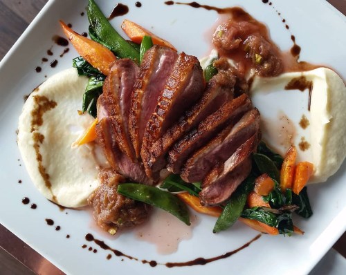 A beautifully plated dish featuring sliced duck breast, creamy puree, sautéed vegetables, and a drizzle of sauce.