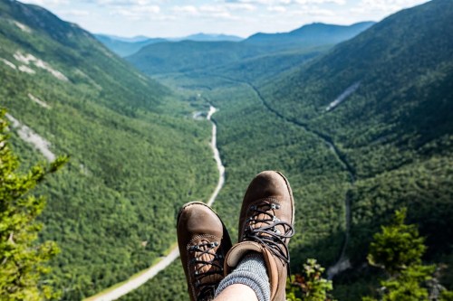 A pair of hiking boots resting on a ledge, overlooking a lush green valley and winding river below.