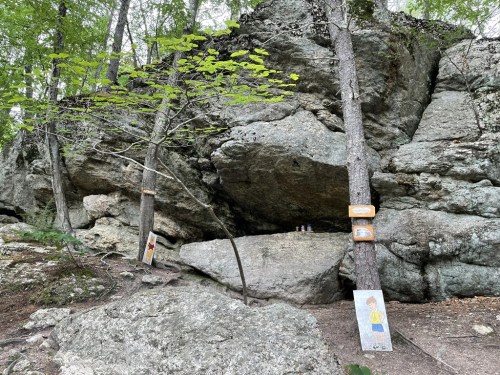 A rocky area in a forest with trees and signs, featuring a large boulder and painted figures on the ground.
