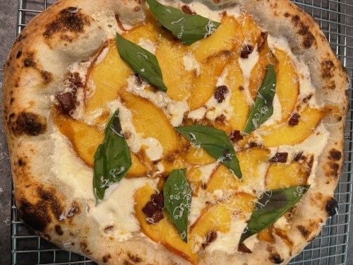 A freshly baked pizza topped with sliced peaches, basil leaves, cheese, and a golden crust on a cooling rack.