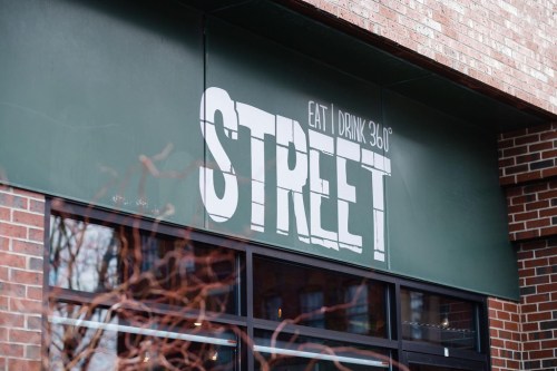 Sign for "STREET" restaurant, featuring the words "EAT | DRINK 360°" on a green background, against a brick wall.
