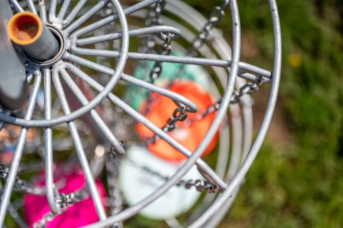 A close-up view of a metal disc golf basket with colorful discs hanging from the chains, set against a grassy background.
