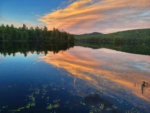 A serene lake at sunset, reflecting colorful clouds and surrounded by lush trees and distant hills.