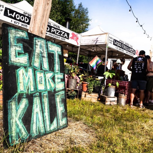 A chalkboard sign reads "Eat More Kale" in front of food tents at an outdoor event, surrounded by plants and people.