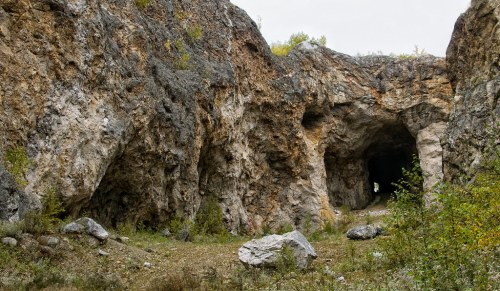 A rocky cave entrance surrounded by rugged terrain and sparse vegetation under a cloudy sky.