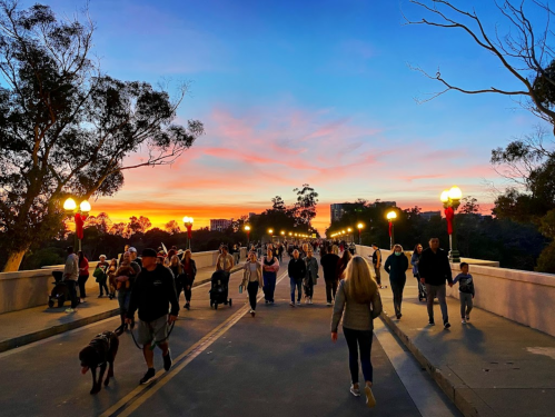 A vibrant sunset casts colors over a busy bridge filled with people walking, some with dogs, surrounded by trees.