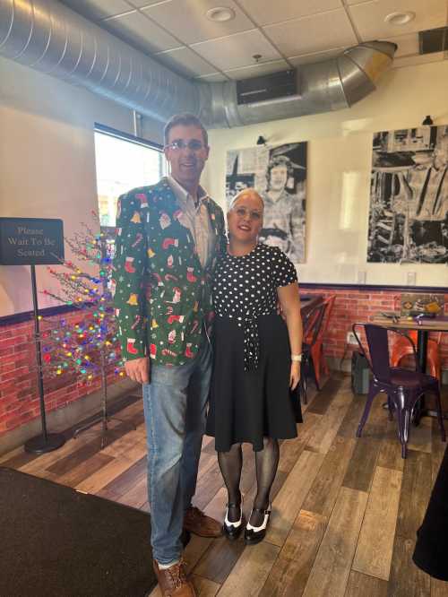 A man in a festive Christmas jacket stands next to a woman in a polka dot dress, both smiling in a decorated restaurant.