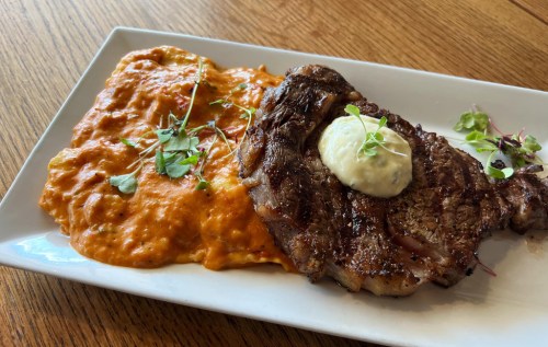 A grilled steak topped with a dollop of sauce, served alongside creamy tomato risotto garnished with microgreens.