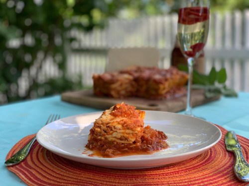 A slice of lasagna on a white plate, garnished with sauce, set on a colorful table with a glass of sparkling drink in the background.