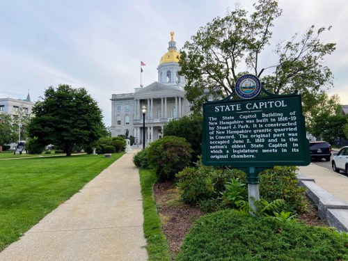 The New Hampshire State Capitol building with a golden dome, surrounded by greenery and a historical sign in the foreground.