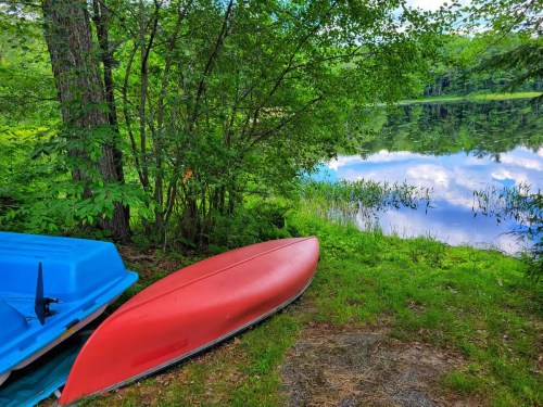 A red kayak and a blue boat are resting on the grass by a calm lake surrounded by lush greenery.