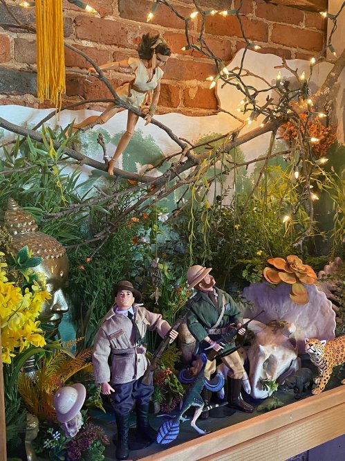 A colorful display featuring action figures, plants, and a leopard statue against a brick wall backdrop.