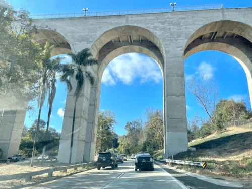 A view of a highway passing under a large concrete bridge with arches, surrounded by trees and a blue sky.
