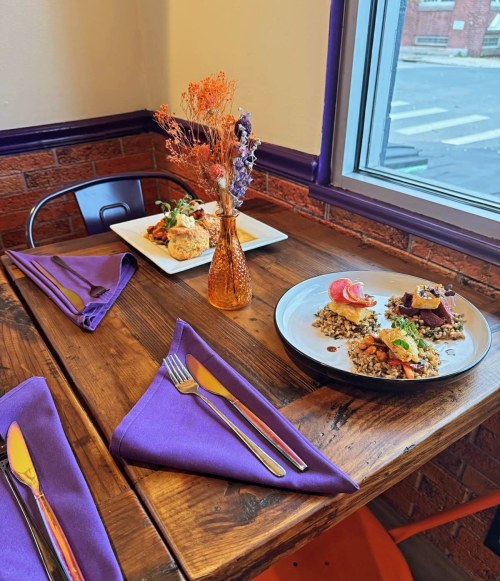 A cozy table setting with two plates of food, a vase of dried flowers, and purple napkins in a warm, inviting space.