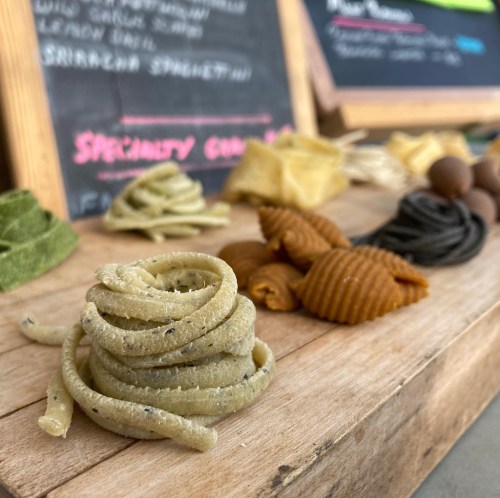 A variety of colorful, artisanal pasta shapes displayed on a wooden surface, with a chalkboard menu in the background.