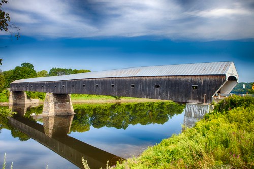 A long, wooden covered bridge spans a calm river, reflecting in the water under a blue sky with scattered clouds.