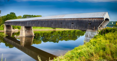 A long, wooden covered bridge spans a calm river, reflecting in the water, surrounded by lush greenery.