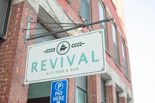 Sign for "Revival Kitchen & Bar" hanging from a brick building, with a parking sign below it.
