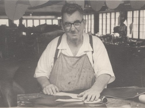 A man in an apron stands at a workbench in a workshop, smiling and looking at the camera.