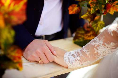 A close-up of two hands clasped together, one wearing a wedding ring, surrounded by autumn leaves.