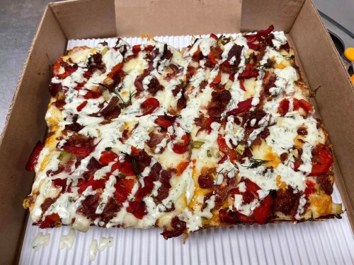 A rectangular pizza topped with bacon, red peppers, and drizzled with white sauce, in a cardboard box.