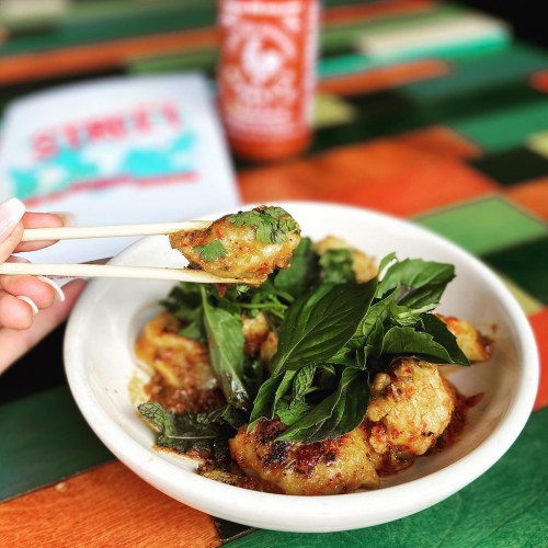 A hand holding chopsticks with a flavorful dish of dumplings topped with fresh herbs on a colorful table.
