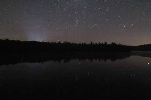 A serene night sky filled with stars reflects on a calm lake, surrounded by dark silhouettes of trees.