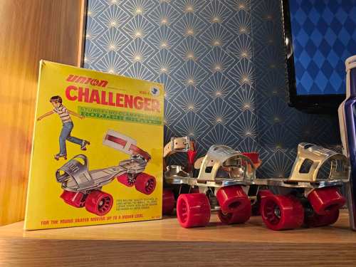 A vintage roller skate box next to a pair of red and silver roller skates on a wooden shelf.