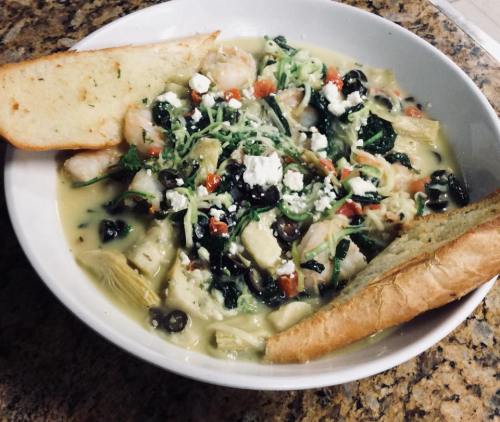 A bowl of creamy pasta with shrimp, spinach, tomatoes, olives, and feta, served with two slices of garlic bread.