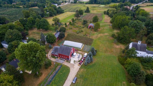 Aerial view of a rural farm with a red barn, green fields, and various crops surrounded by trees and houses.