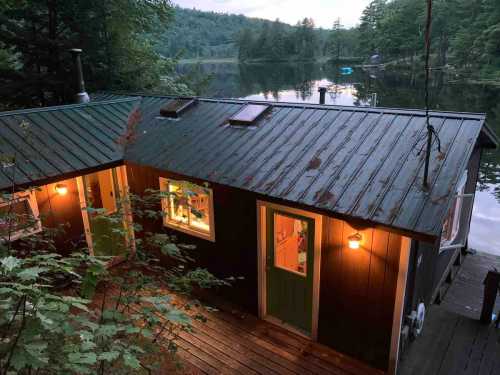 A cozy cabin by a serene lake, surrounded by trees, with warm lights glowing from the windows at dusk.