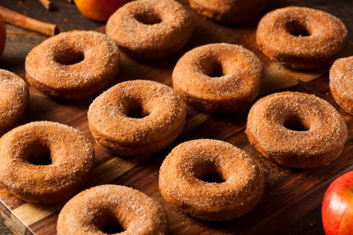 A wooden platter filled with freshly baked cinnamon sugar donuts, surrounded by apples and cinnamon sticks.