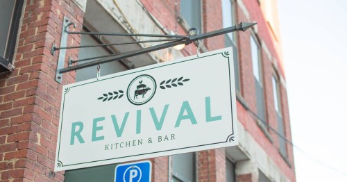 Sign for "Revival Kitchen & Bar" hanging from a brick building, featuring a cow graphic and decorative elements.