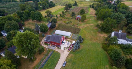 Aerial view of a rural farm with a red barn, green fields, and various crops surrounded by trees and houses.