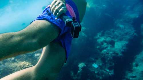 A person swimming underwater, holding a camera, with colorful coral visible in the background.