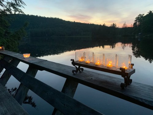 A serene lakeside view at sunset, featuring glowing candles on a wooden railing with mountains in the background.