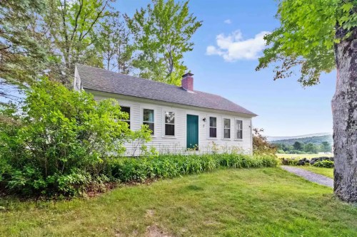 A charming white cottage surrounded by greenery and trees, with a blue door and a scenic landscape in the background.
