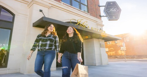 Two women walk outside a restaurant, smiling and carrying a shopping bag, with sunlight illuminating the scene.