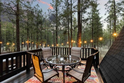 A cozy outdoor deck with a round table and chairs, surrounded by tall pine trees and string lights at dusk.