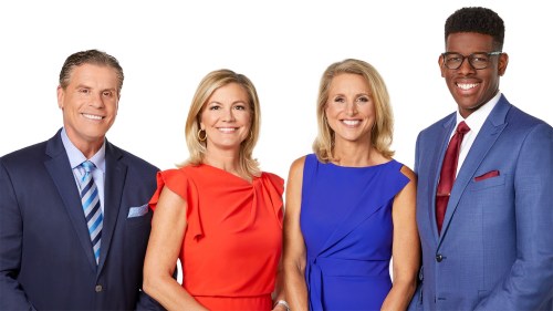 Four smiling news anchors pose together, dressed in professional attire against a white background.