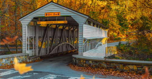 A white covered bridge surrounded by autumn foliage, with a stone wall and road leading to it.